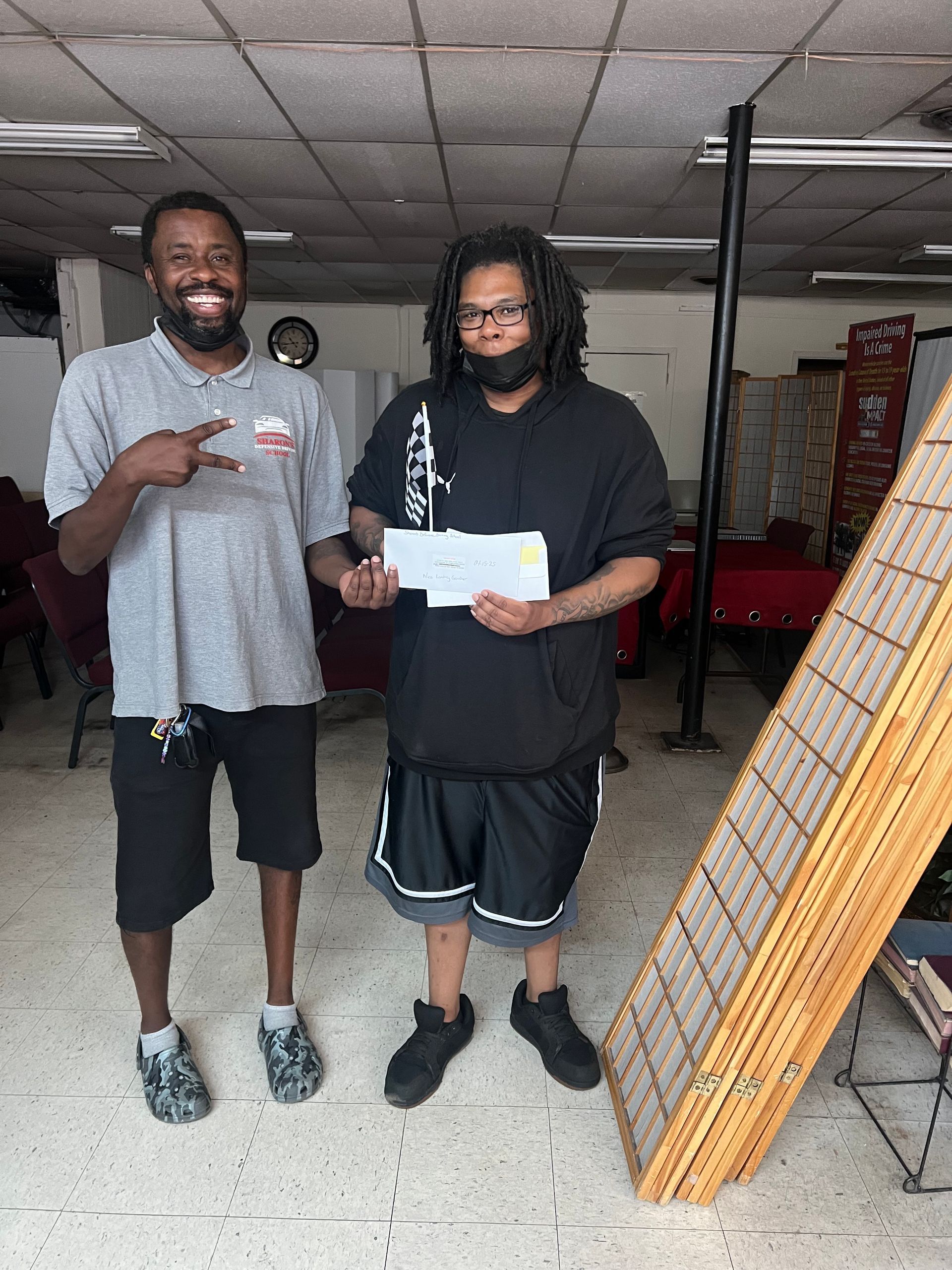 Two men standing inside a doorway, one holds a check, the other gestures a peace sign.