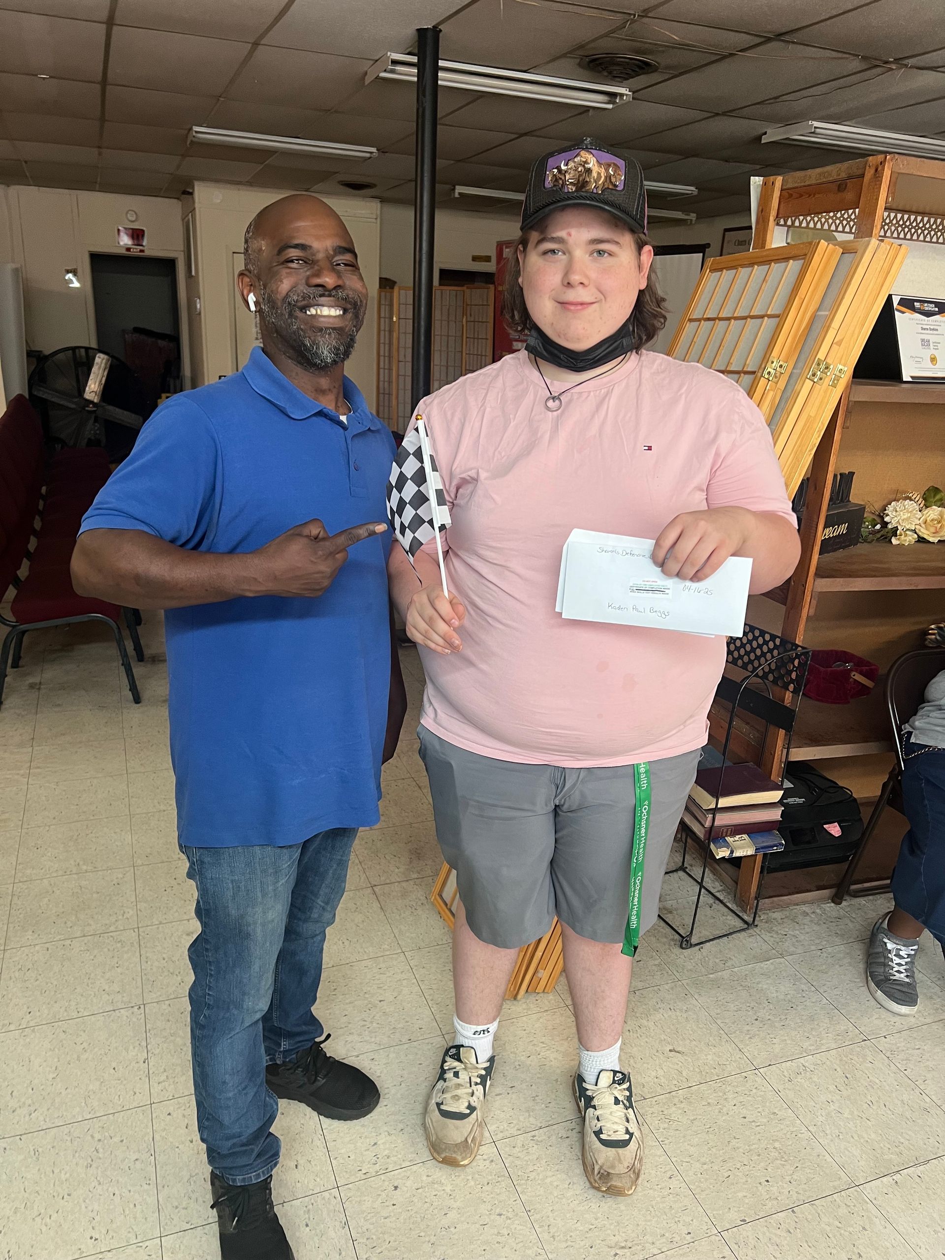 Man in blue shirt points at person holding a paper; both smile. They stand inside a room with shelves.