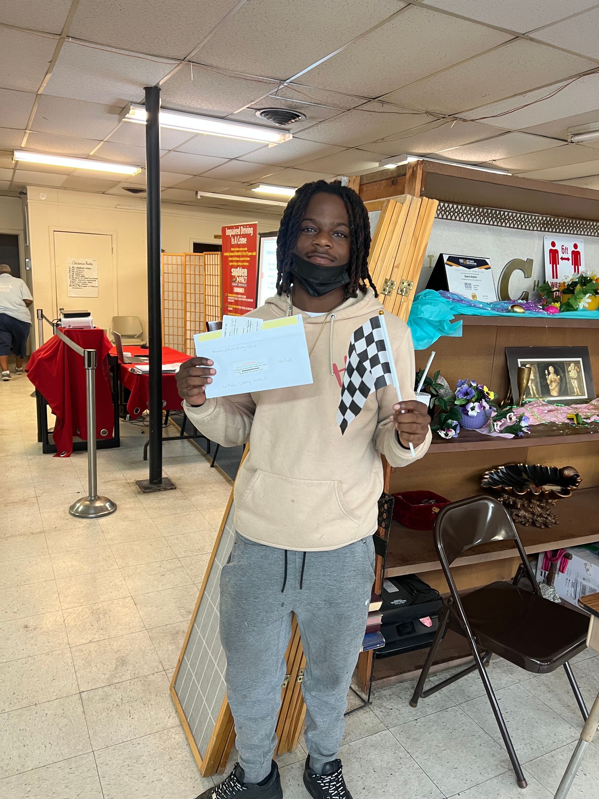 Person holding papers, smiling, in a shop. Wearing light hoodie, grey pants, black shoes and face mask.