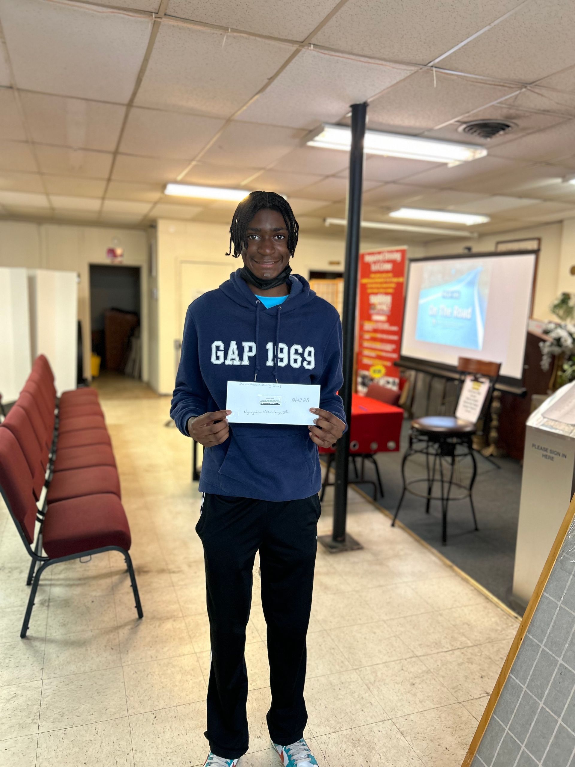 Teenager in blue hoodie holds a paper, standing inside a room with chairs and a screen.