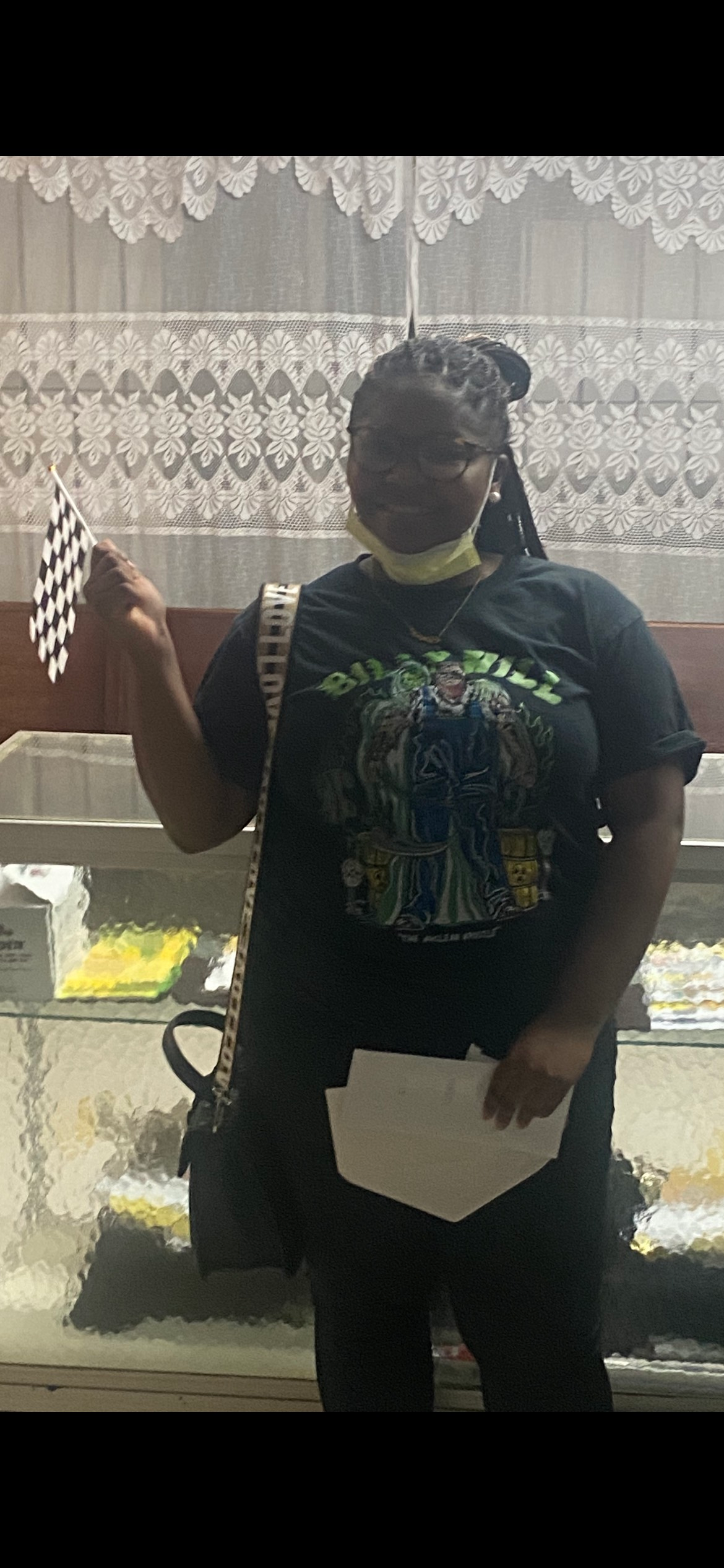 Woman holding a small black and white checkered flag and a paper, standing in front of a white lace curtain.