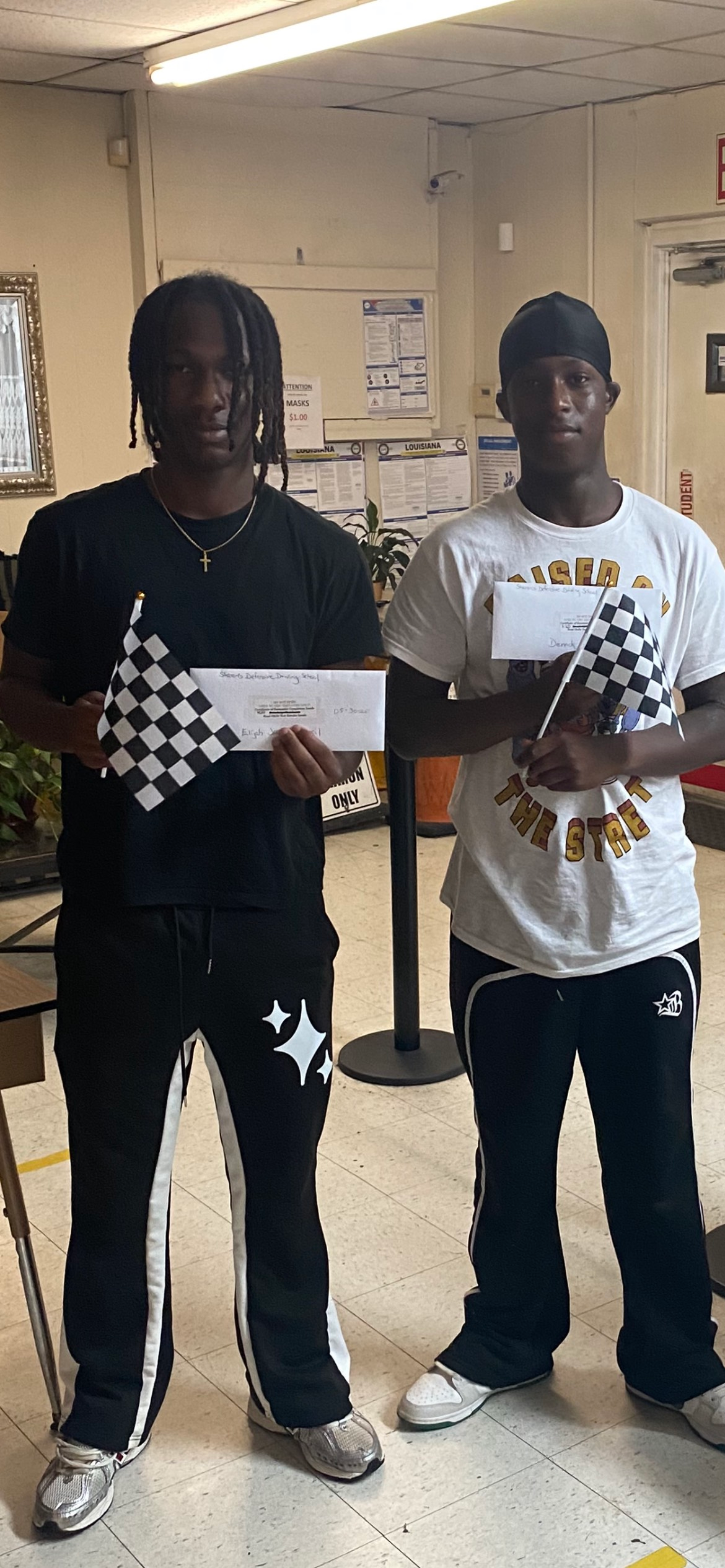 Two people hold checkered flags and envelopes, smiling. Indoor setting.