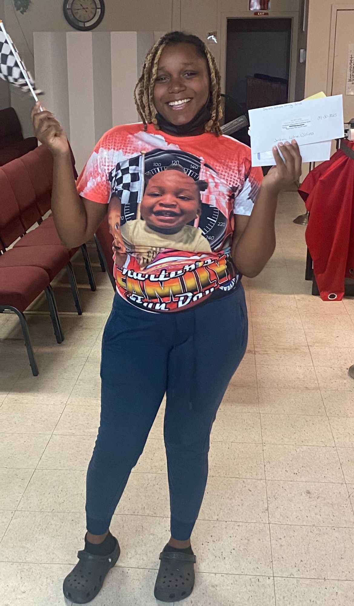 Woman holding a checkered flag and a paper, smiling. Wearing a shirt with a baby's image and blue pants, in a room with pews.