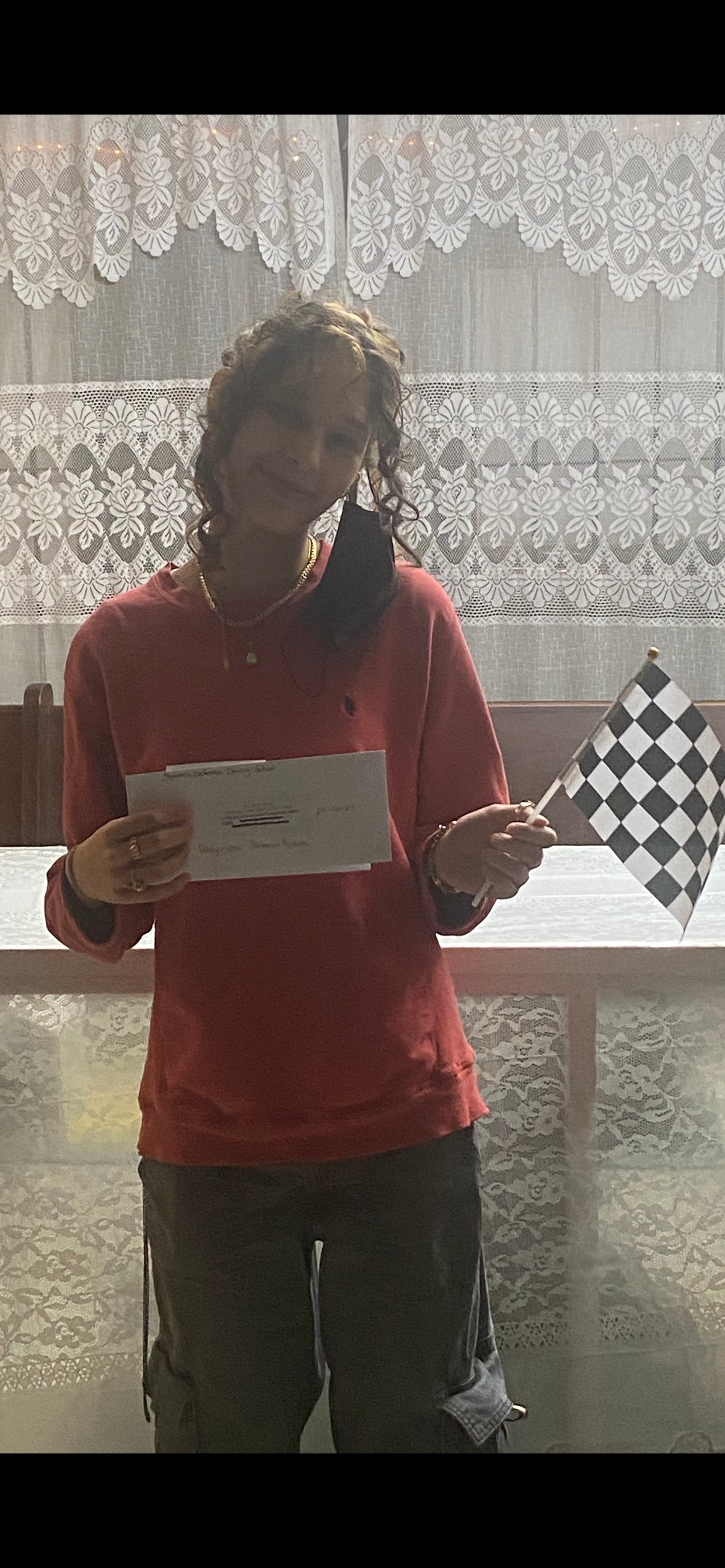 Person in red shirt holding a white paper and a checkered flag, smiling indoors.