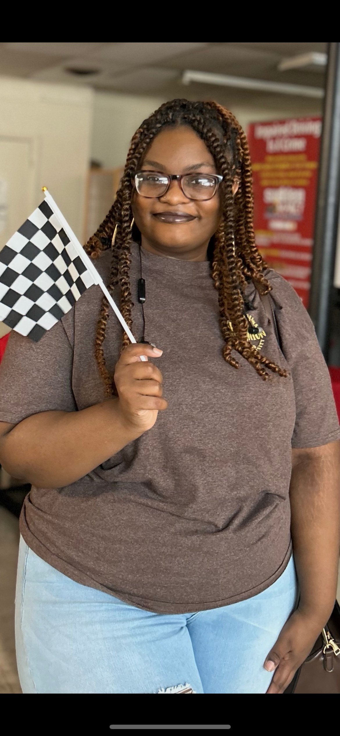 Person holding a checkered flag, smiling, brown shirt, blue jeans, braids, glasses.