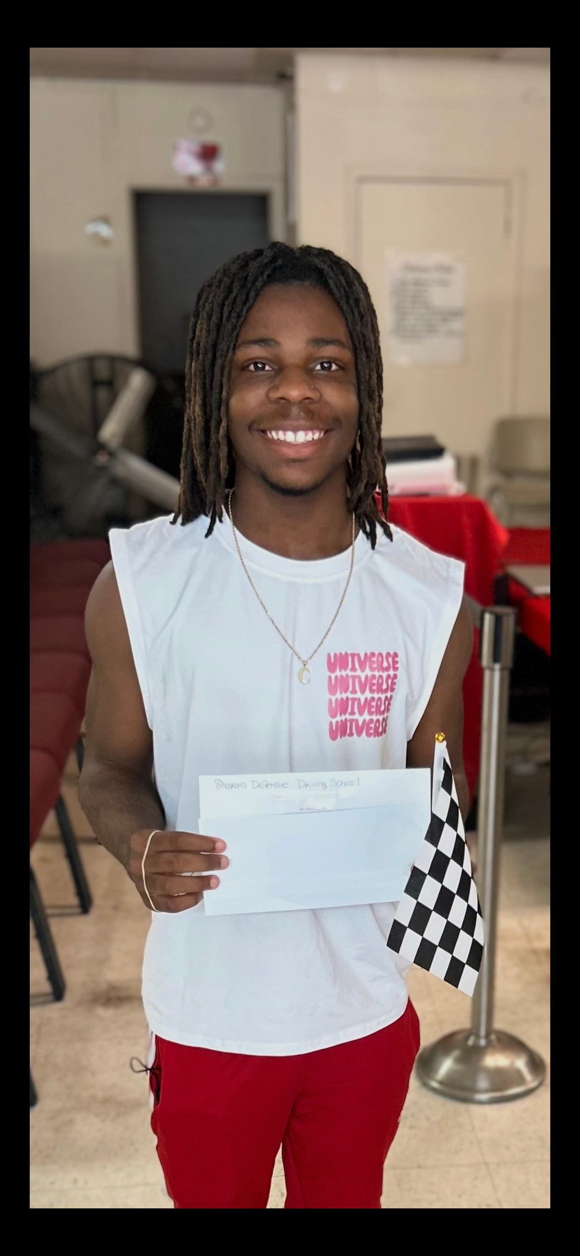 Person holding paper and checkered flag, smiling. Wearing white sleeveless shirt and red pants. Indoors.