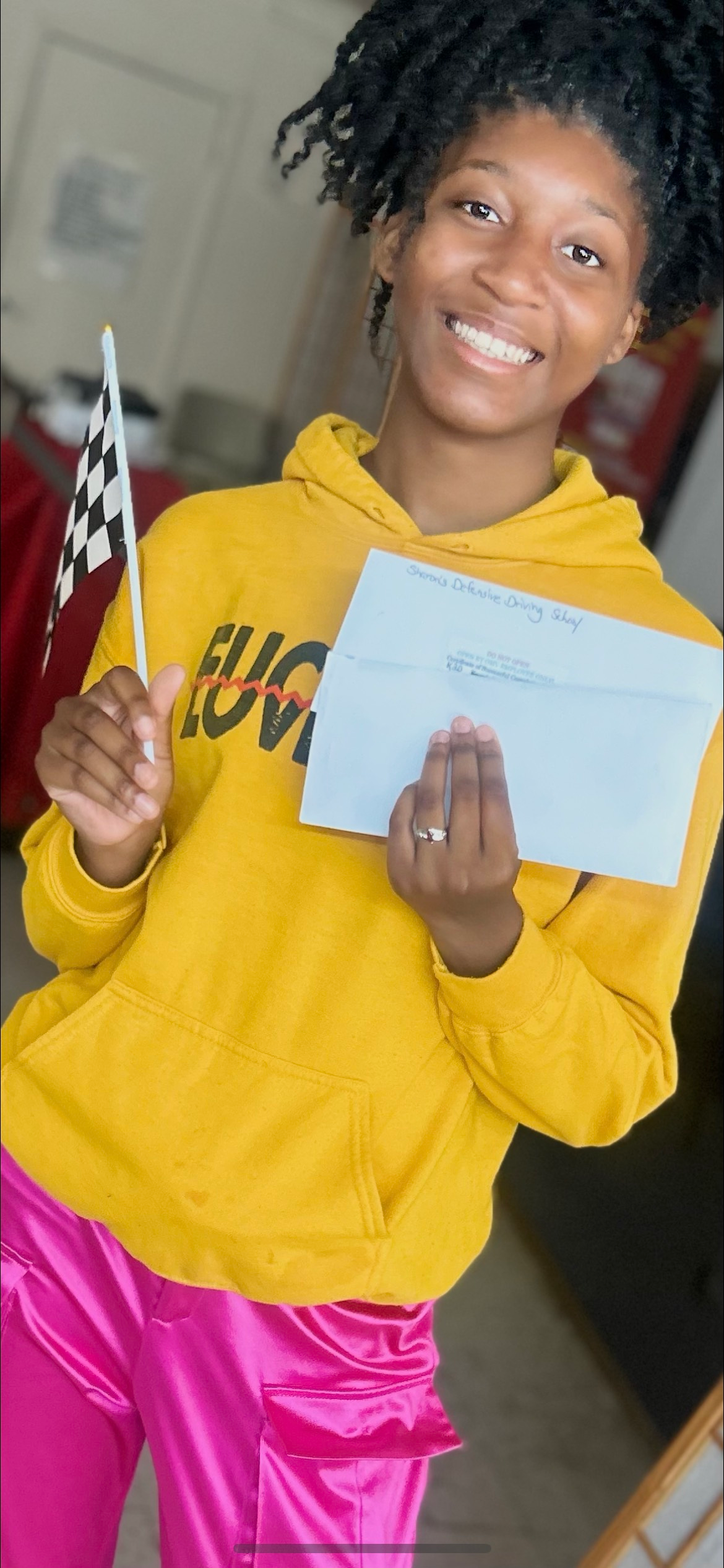 Person in yellow hoodie and pink pants holding a checkered flag and document, smiling.