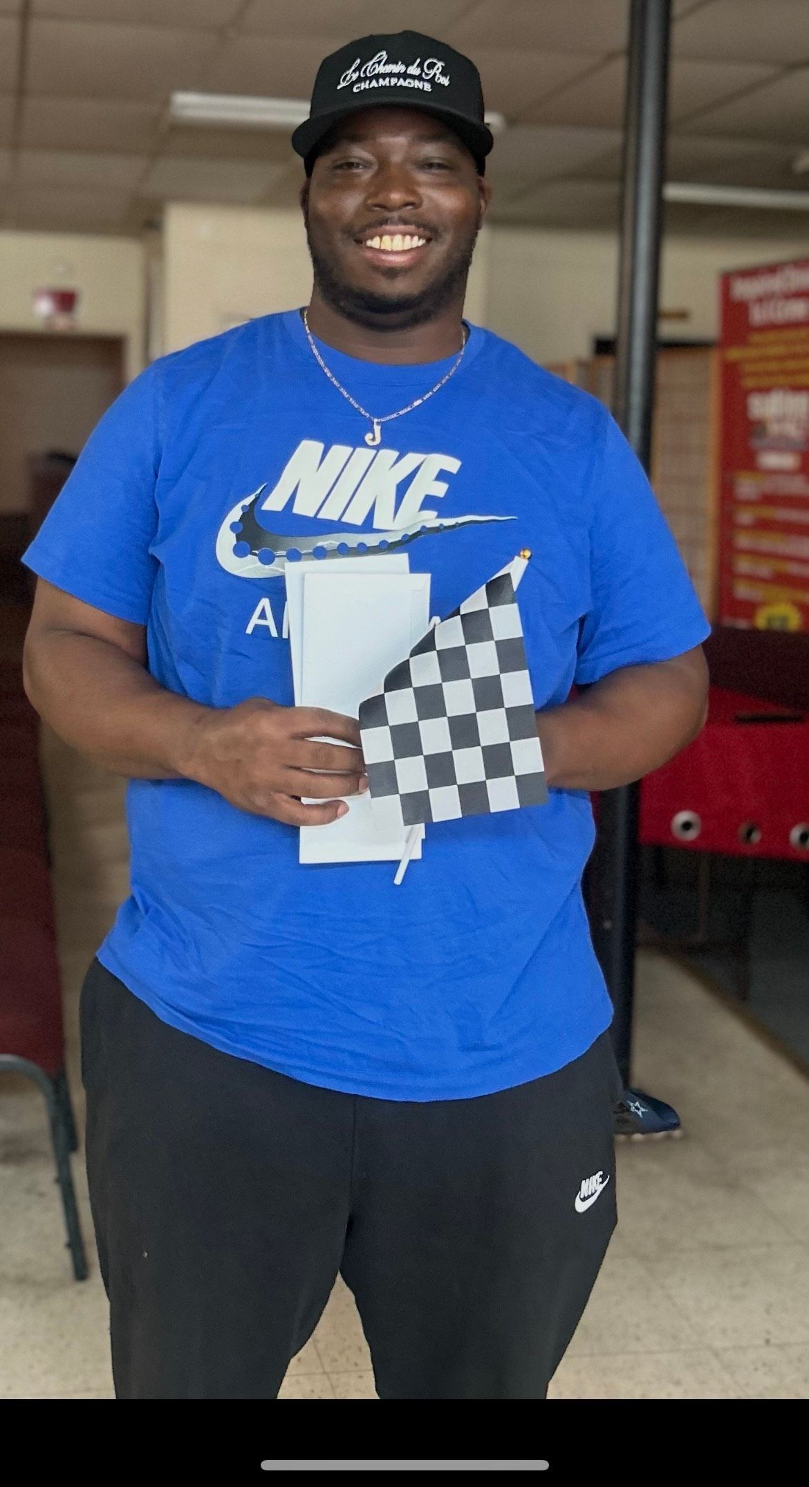 Man in blue Nike shirt holding a checkered flag and a paper, smiling.