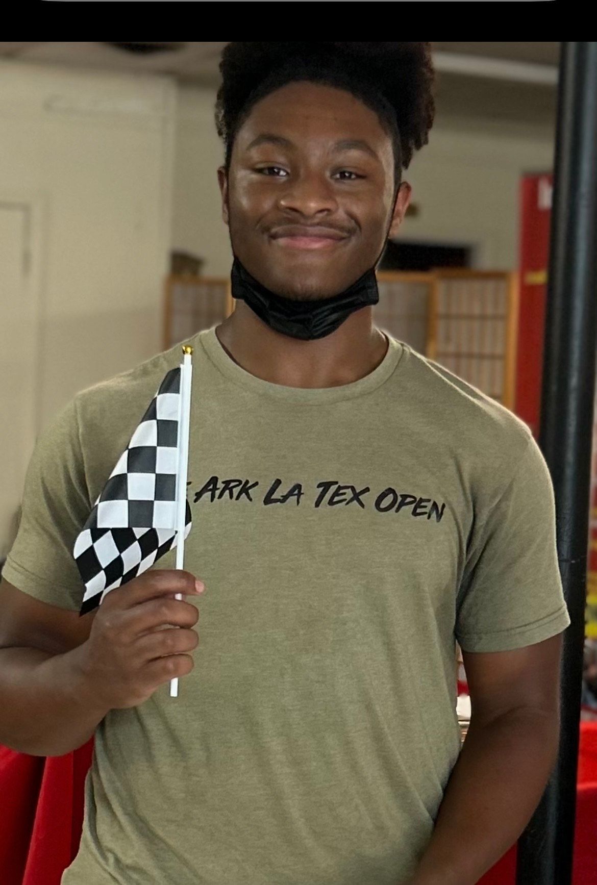 Man wearing a face mask, smiles, and holds a checkered flag. He wears an olive green shirt that reads 