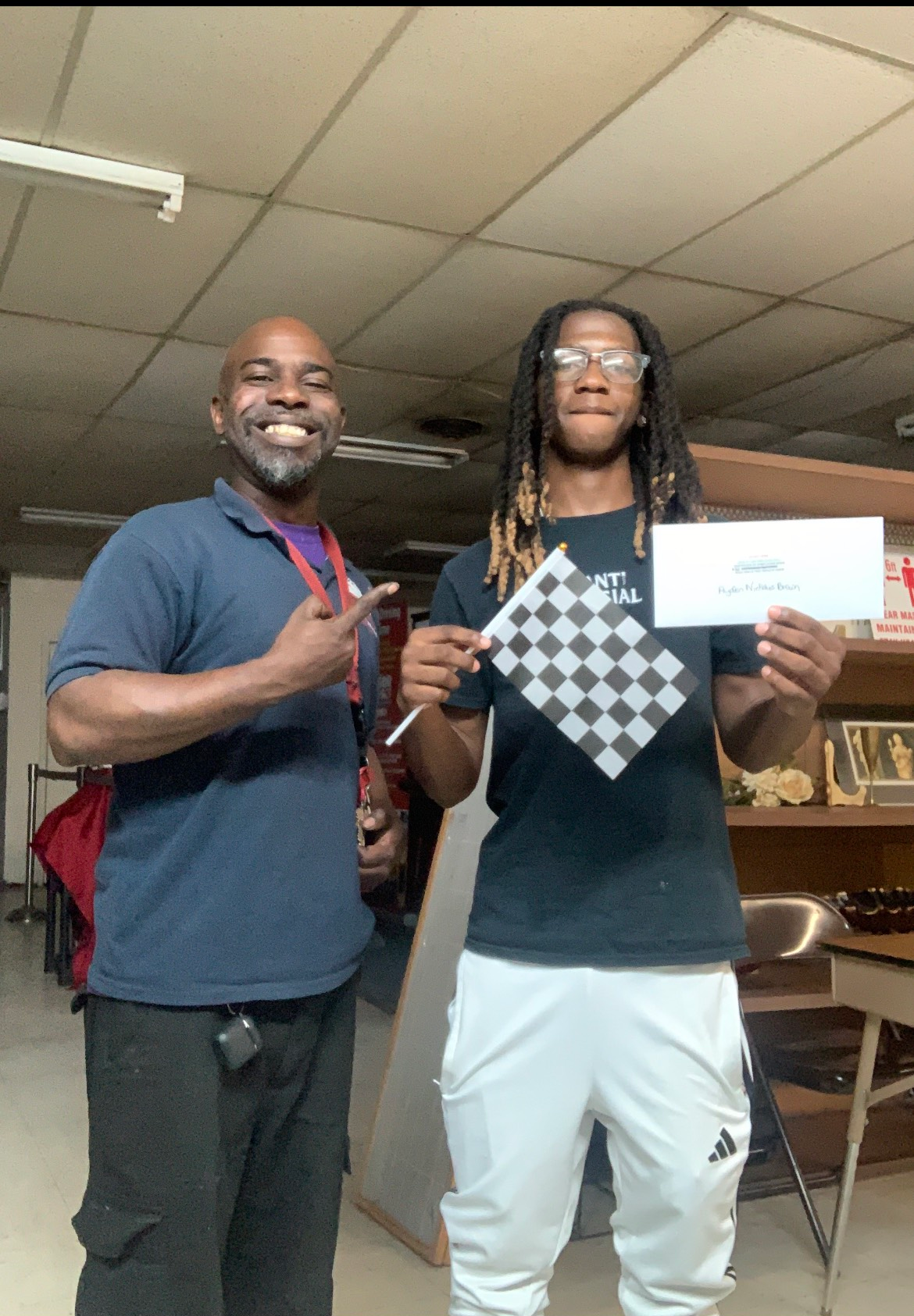 Man points to teen holding checkered flag and certificate. Indoor setting.
