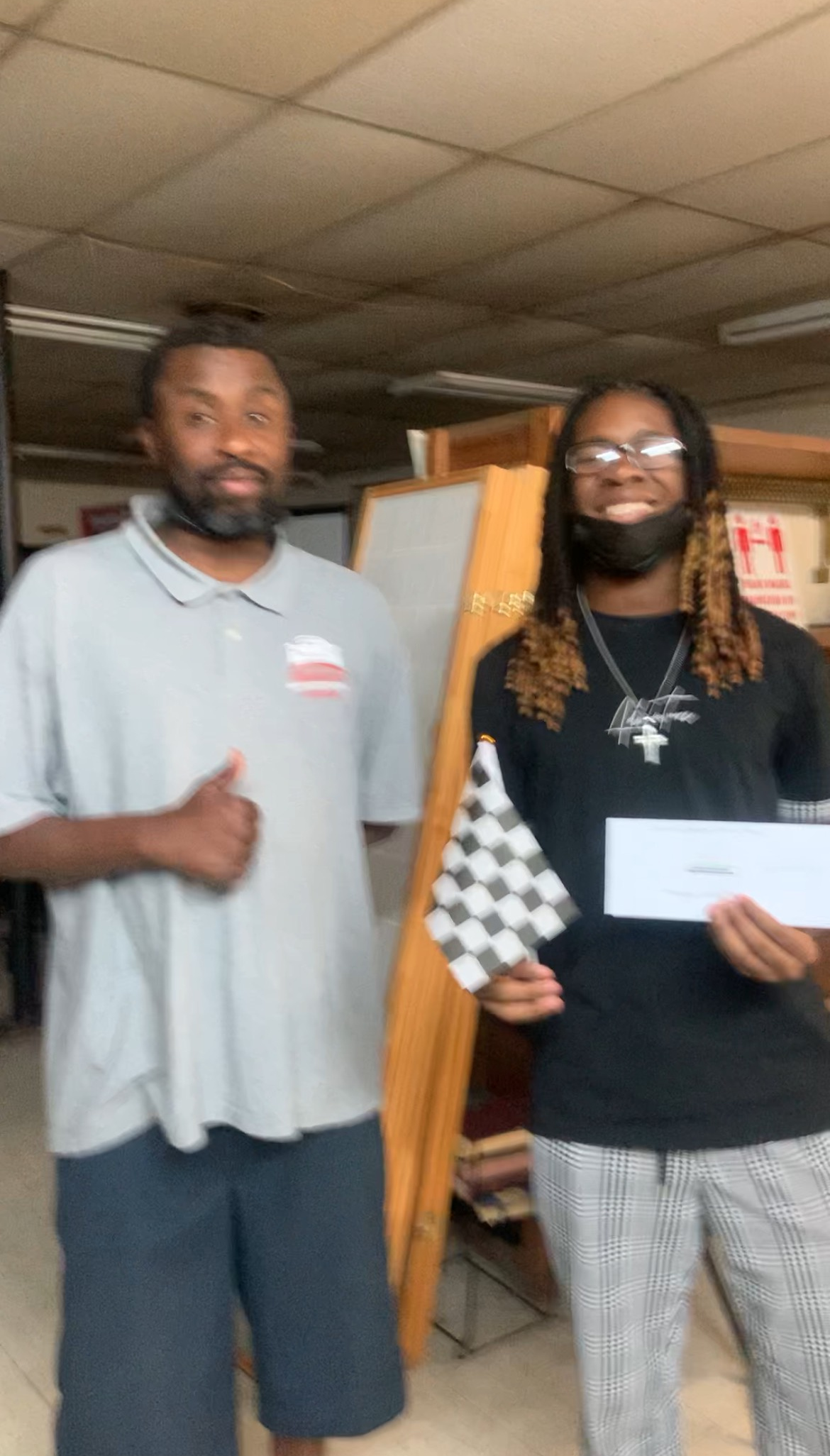 Two men, one holding a checkered flag and document, smiling. One gives a thumbs-up. Indoors, office setting.