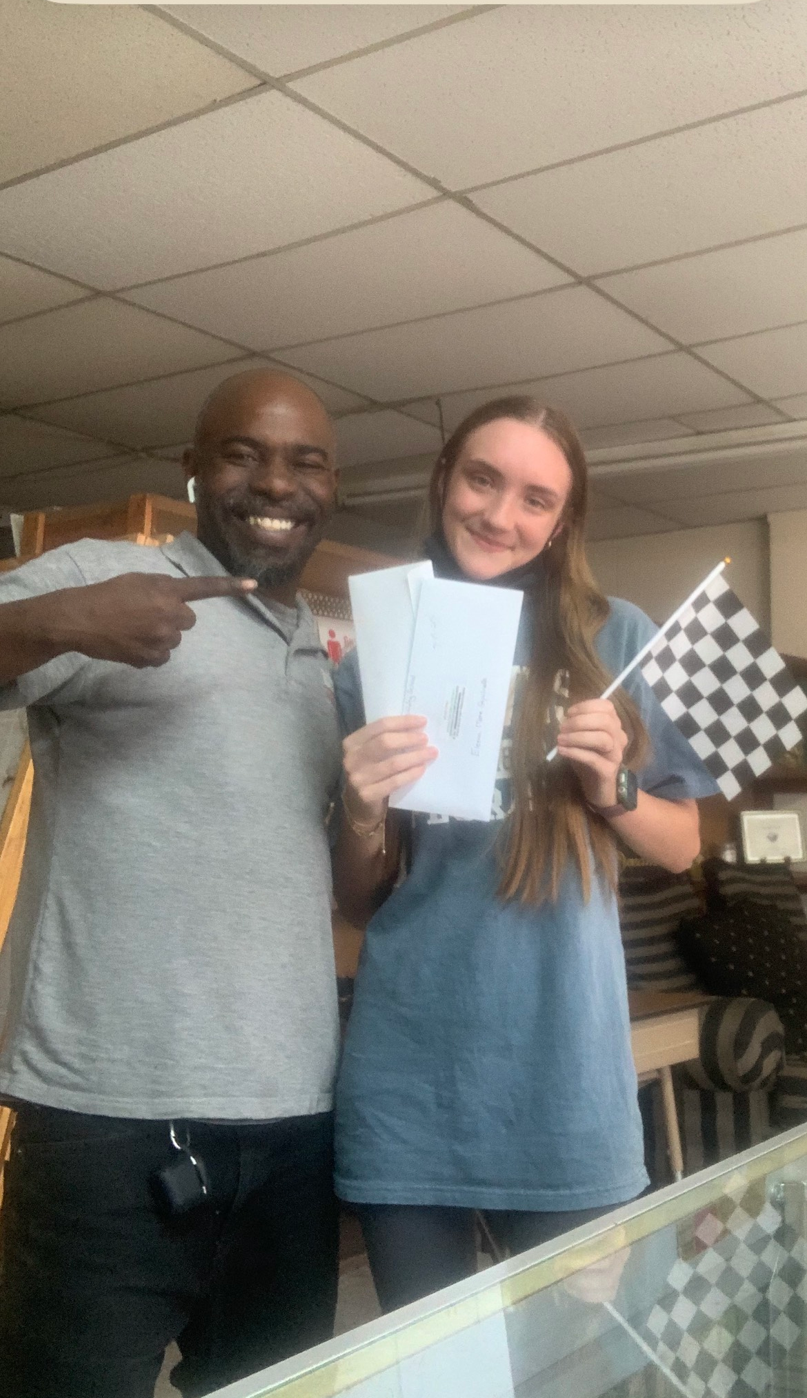 Man points to woman holding paperwork, a checkered flag. Both smile. Indoor setting.