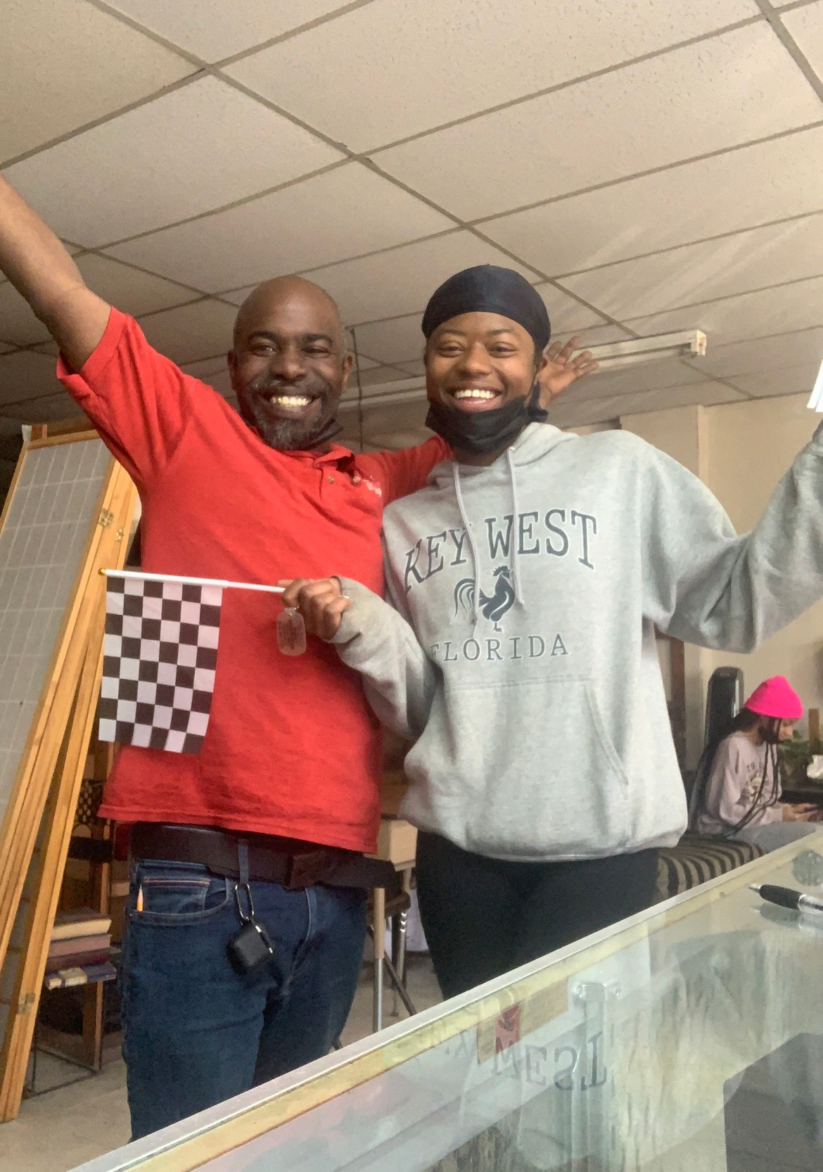 Man and person in gray sweatshirt smiling, holding a checkered flag and raised hand, indoors.