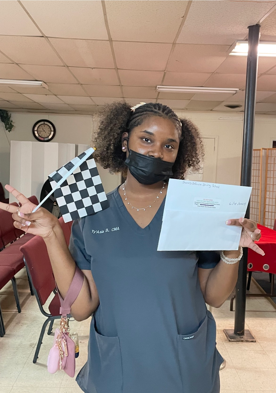Woman in scrubs holds a checkered flag and paper, giving a peace sign, smiling. Interior setting.
