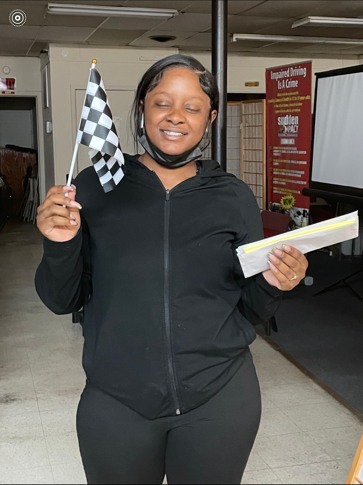 Woman in black outfit holds a checkered flag and a paper, smiling indoors.