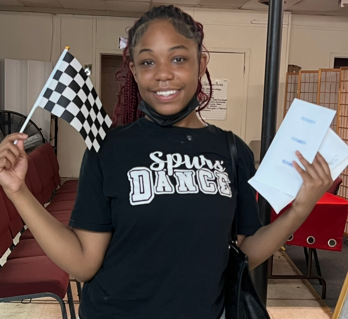 Teenager in black shirt holding a checkered flag and papers, smiling, in a room.