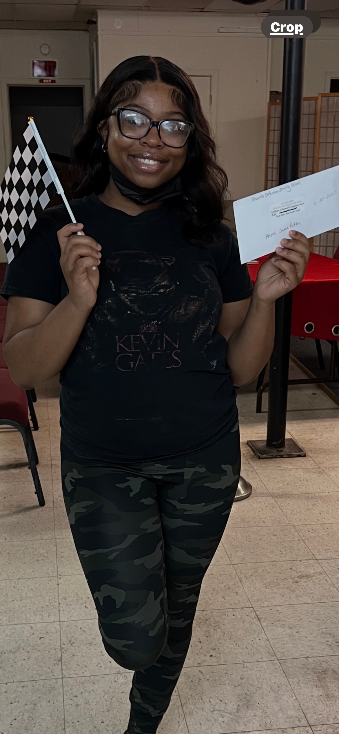 Person holding a checkered flag and a paper, smiling. Wearing black shirt, camo pants, and glasses. Indoors.