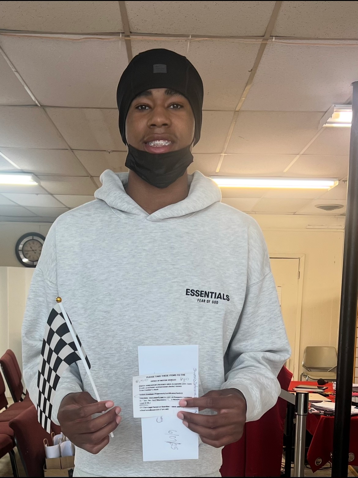 Person in gray hoodie holding a checkered flag and document, smiling. Wearing a black beanie and mask. Indoors.