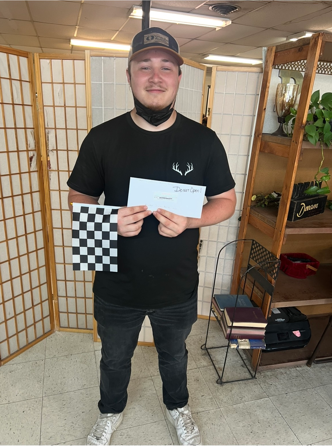 Man in black shirt and hat holds a checkered flag and document, smiling. Indoor setting with dividers and shelves.