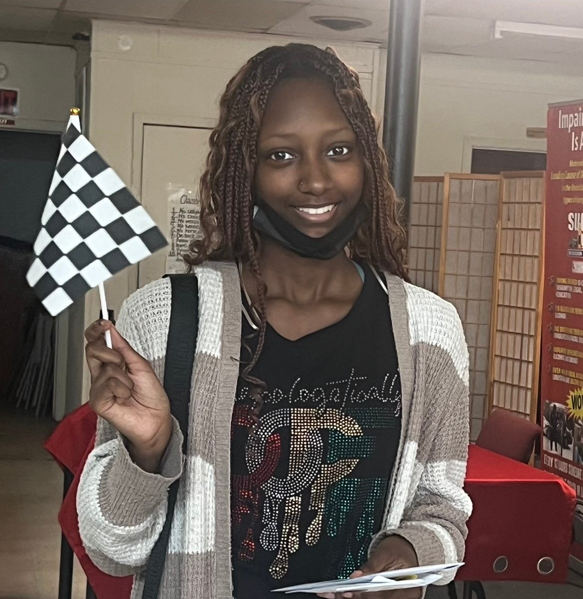 Woman holding a checkered flag, smiling, wearing a mask and cardigan. Interior setting.