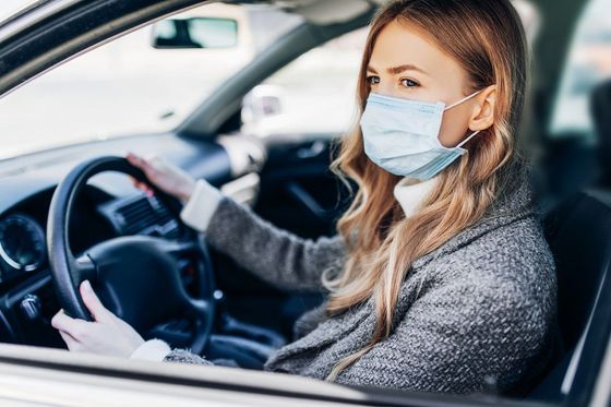 Woman wearing a face mask drives a car, looking out the window, hands on the steering wheel.