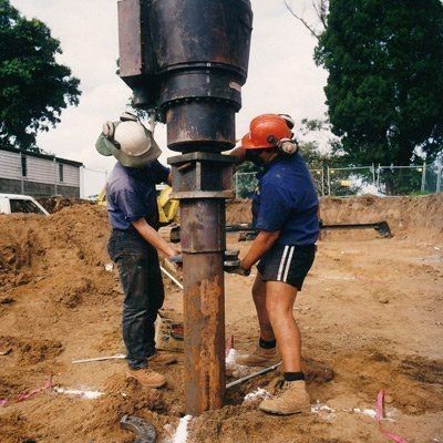 Two construction workers wearing safety gear are performing screw piling at a construction site.