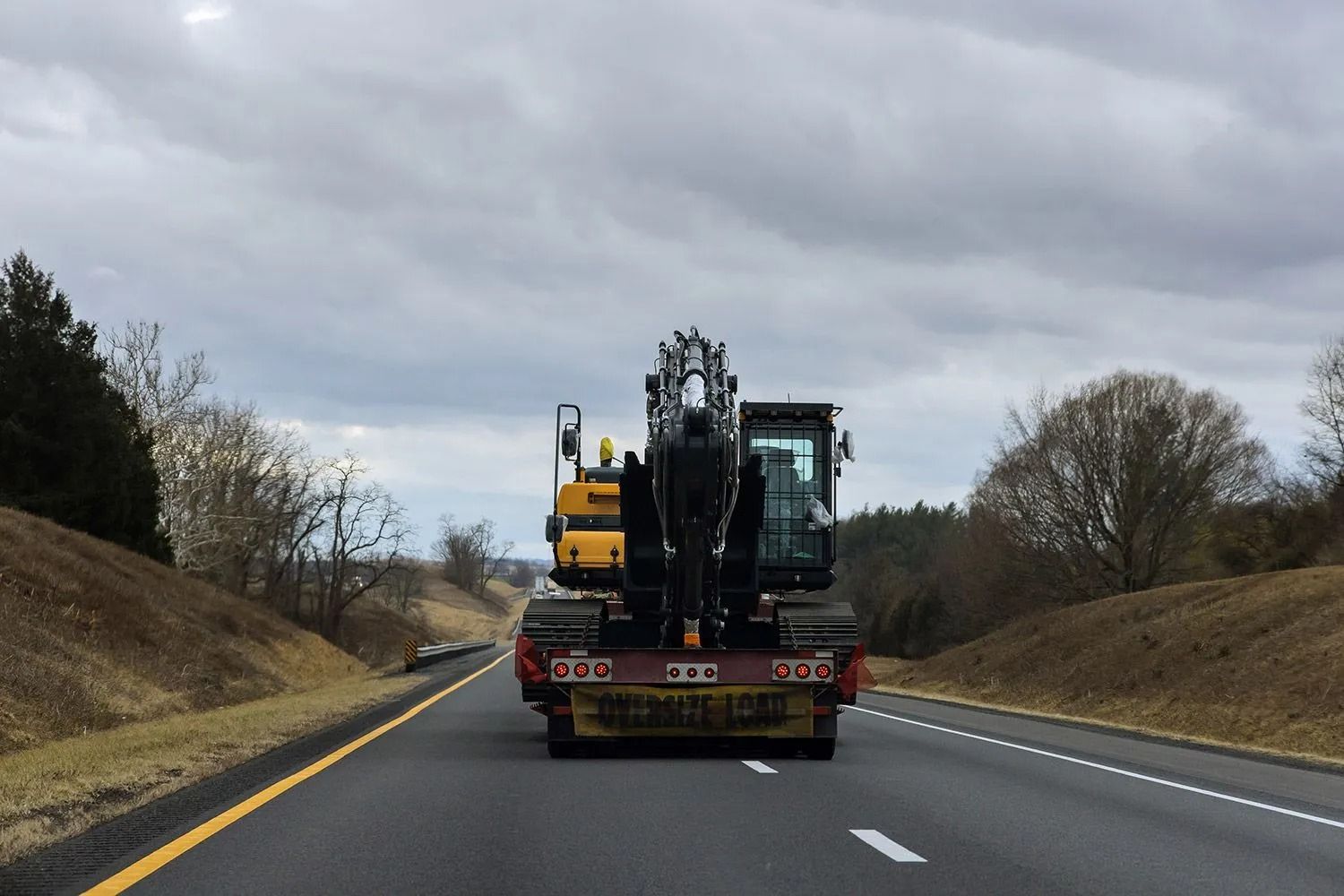 Flatbed truck carrying construction machinery driving on a highway under cloudy skies.