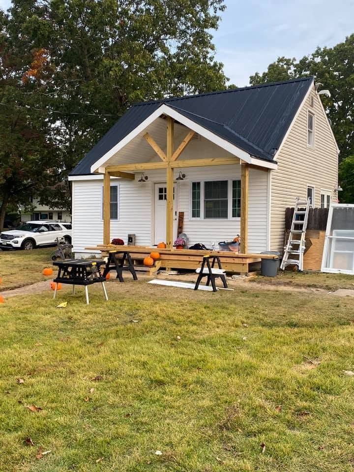 Small white house with black roof, porch posts, and outdoor seating on a grassy yard.