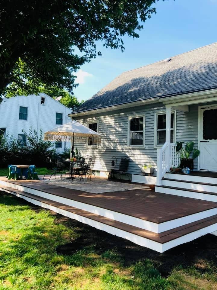 White house with a large brown wooden deck, porch chairs, and a green yard in sunny daylight