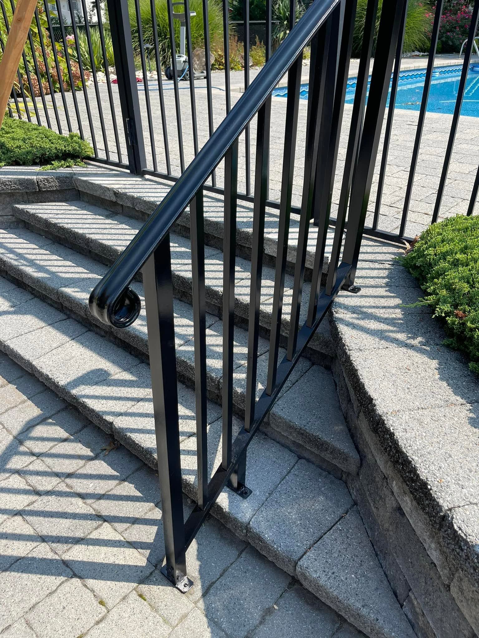 Black metal stair railing beside steps and a path near a pool, with sunlight casting shadows