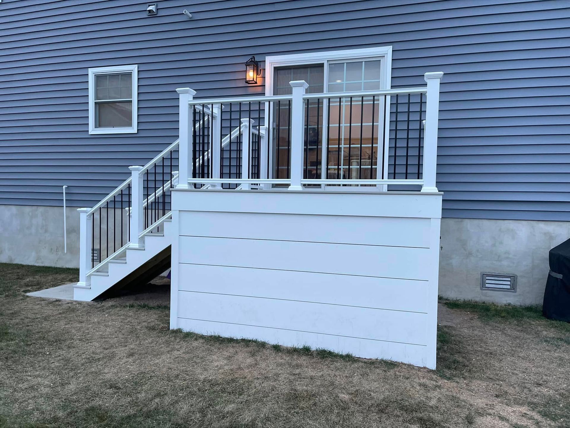 Back deck with white railing and stairs on a blue-sided house, lit by a porch light.