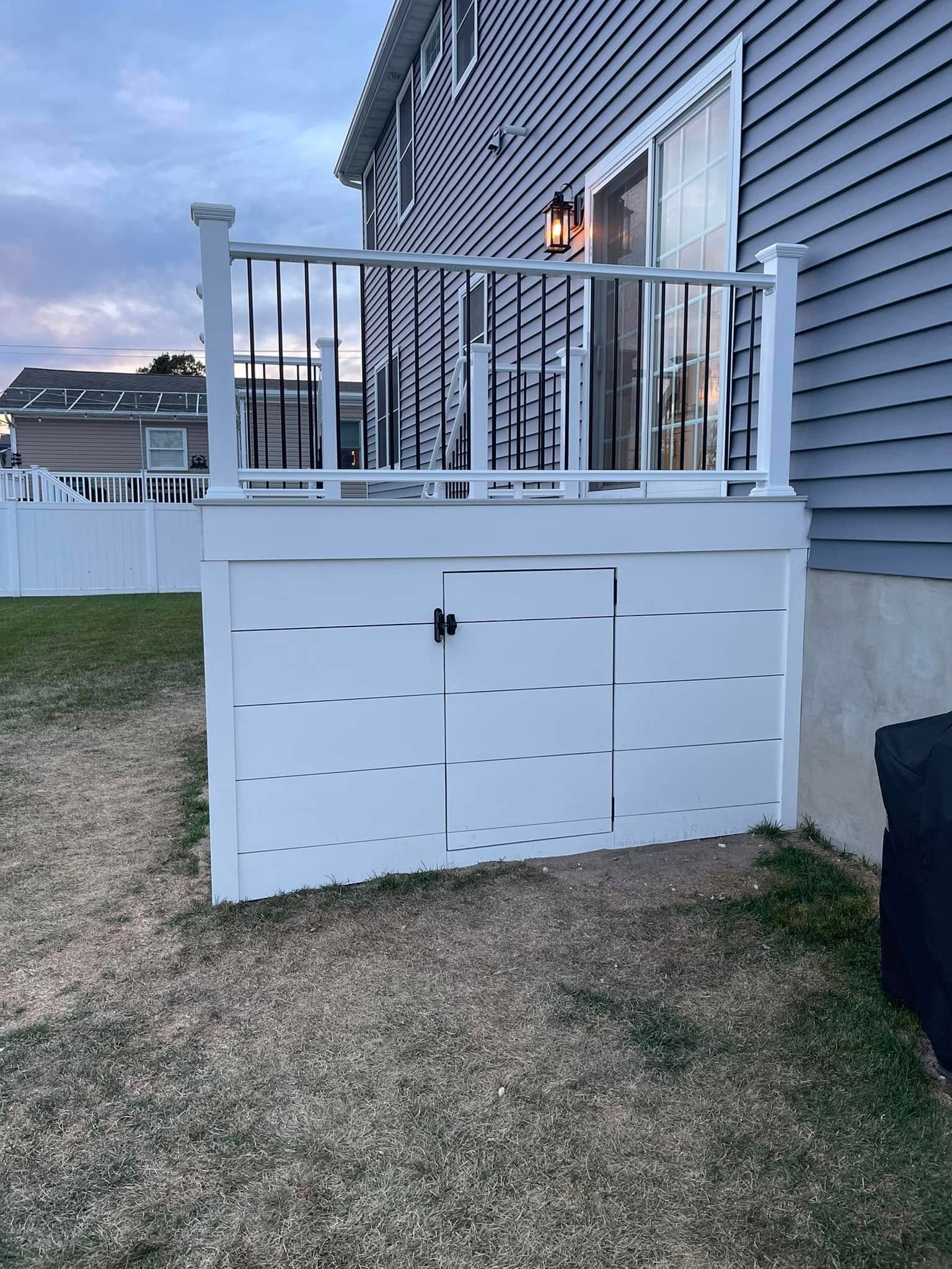 White deck storage enclosure under a small balcony beside a house, with a lit window and grass yard nearby