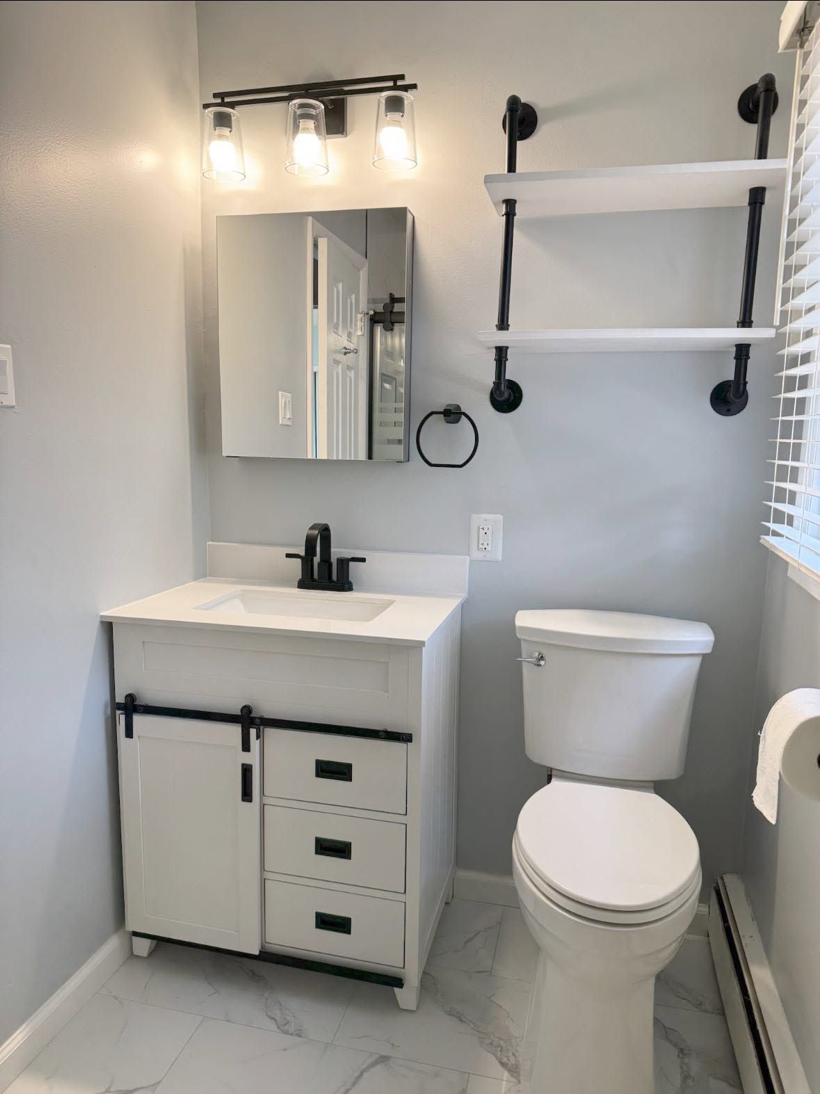 Small bathroom with white vanity, mirror, toilet, black fixtures, wall shelves, and window blinds