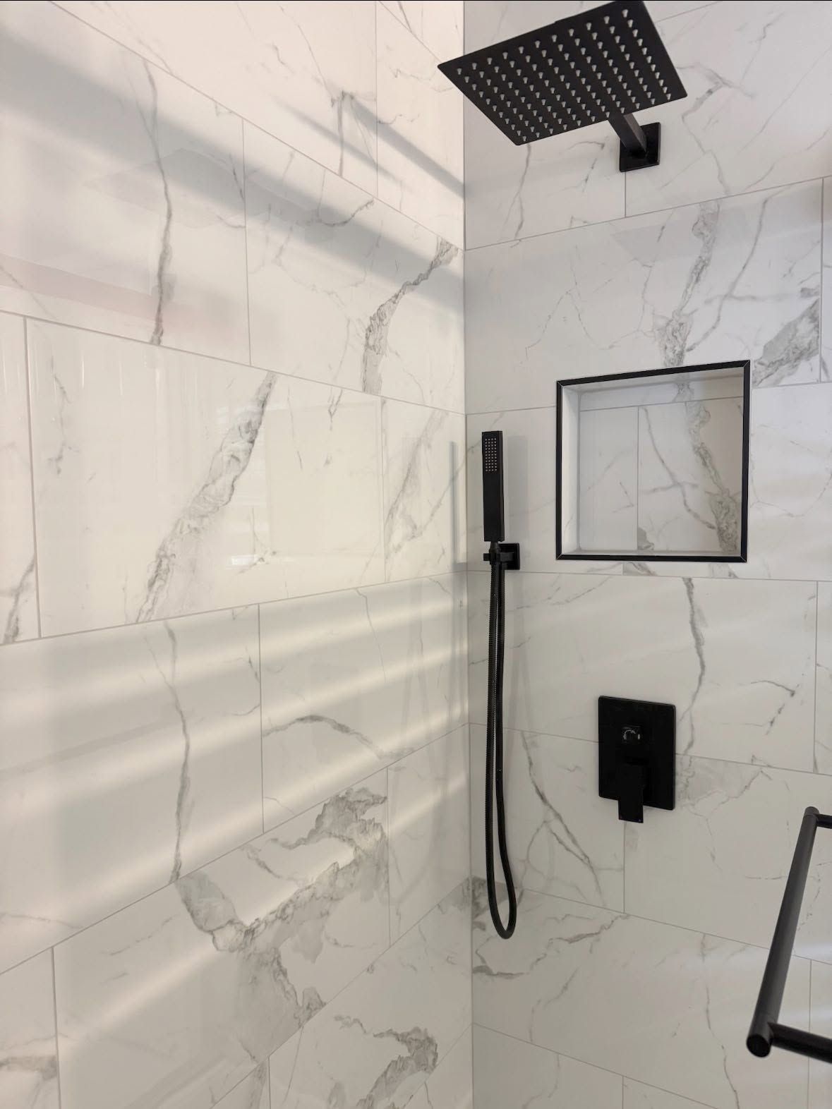 Modern shower with white marble-look tile, black showerhead, handheld wand, and controls