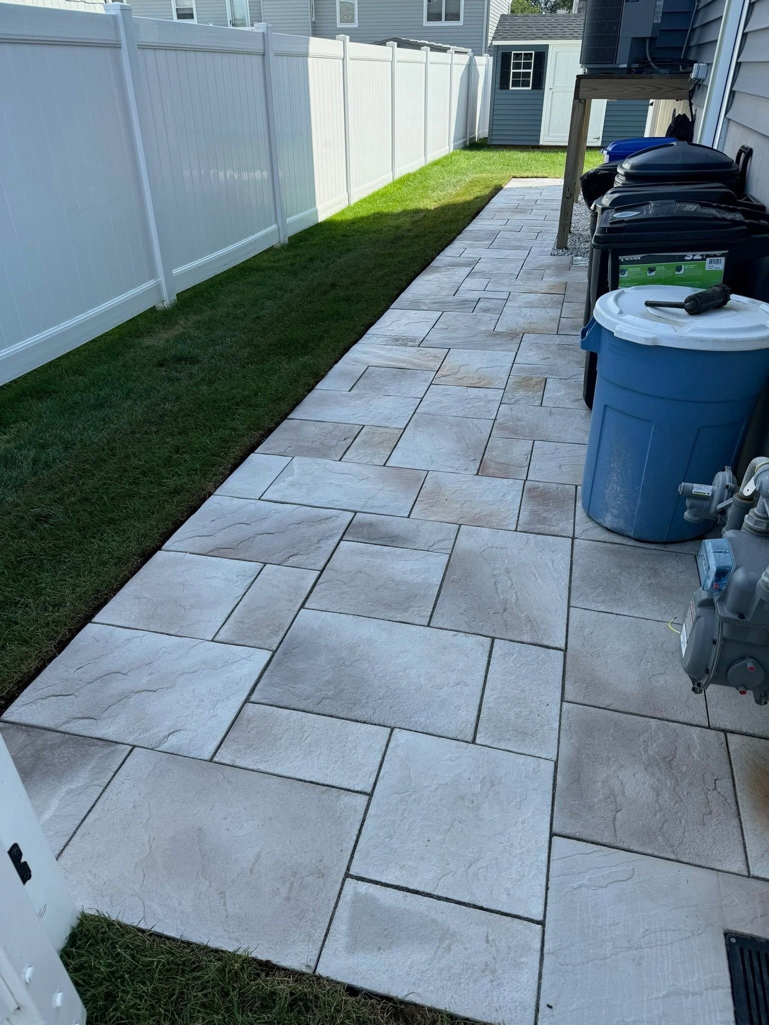 Narrow side yard with a stone walkway, white fence, grass strip, and trash bins along the house.
