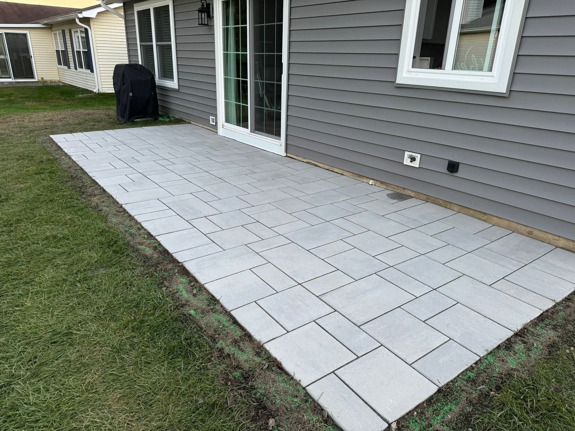 Backyard patio with light gray pavers beside a gray house and lawn