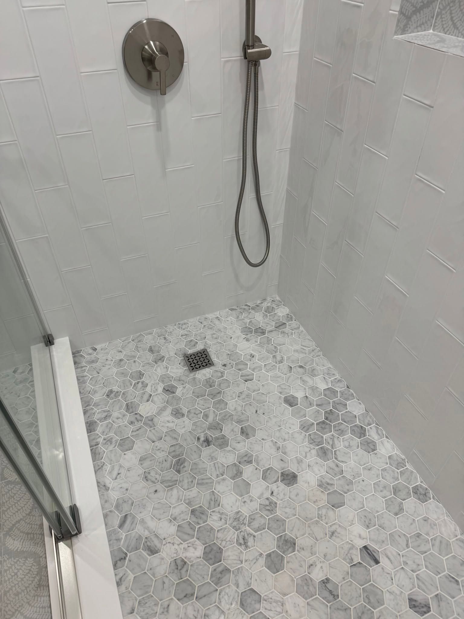 Small white-tiled shower with mosaic floor, handheld showerhead, and glass door side panel.