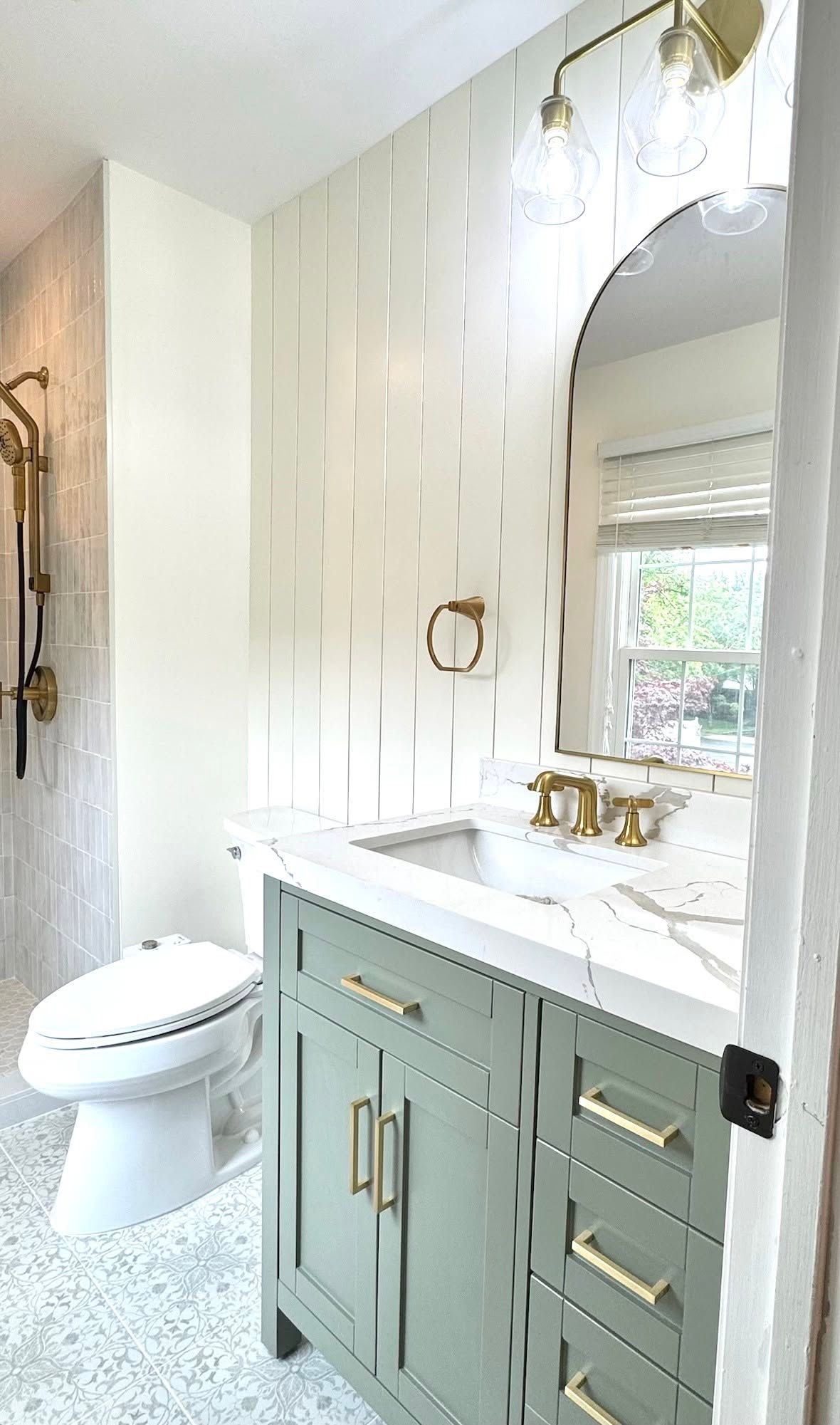 Bright bathroom with green vanity, gold fixtures, oval mirror, and shower curtain by a window.