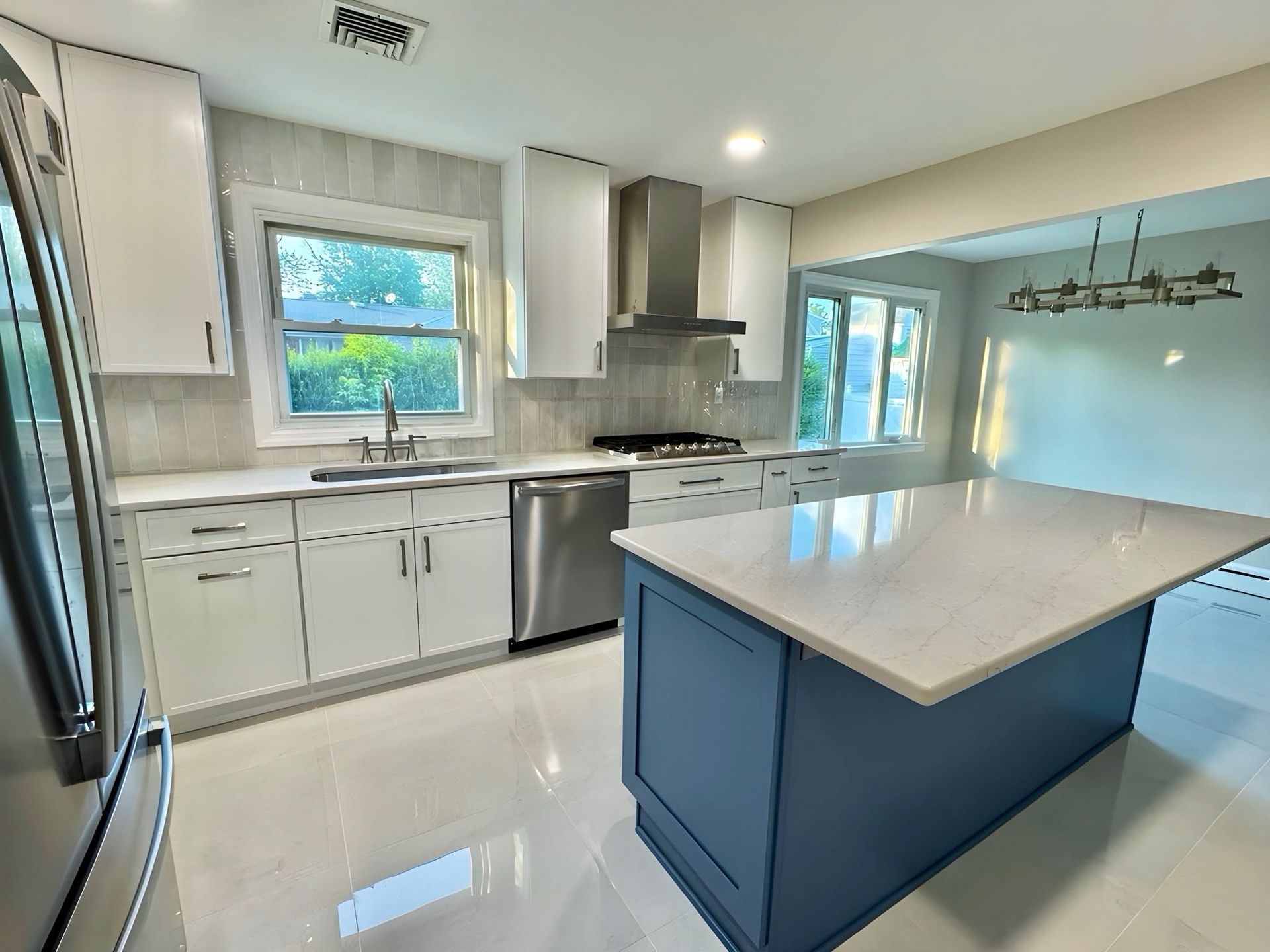 Bright modern kitchen with white cabinets, stainless appliances, and a blue island under large windows.
