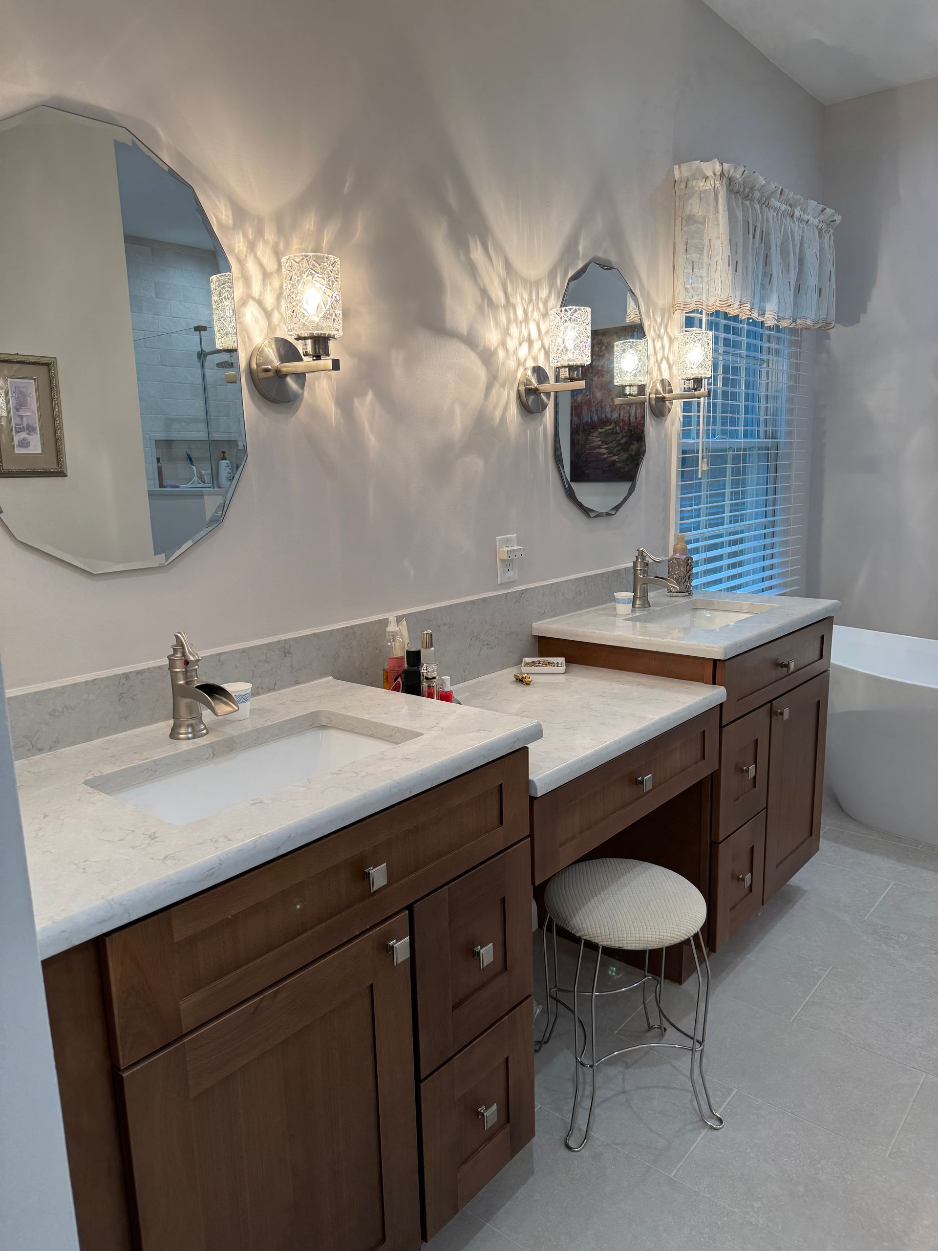 Bright bathroom with double sinks, dark wood vanities, wall sconces, and a window with white blinds.