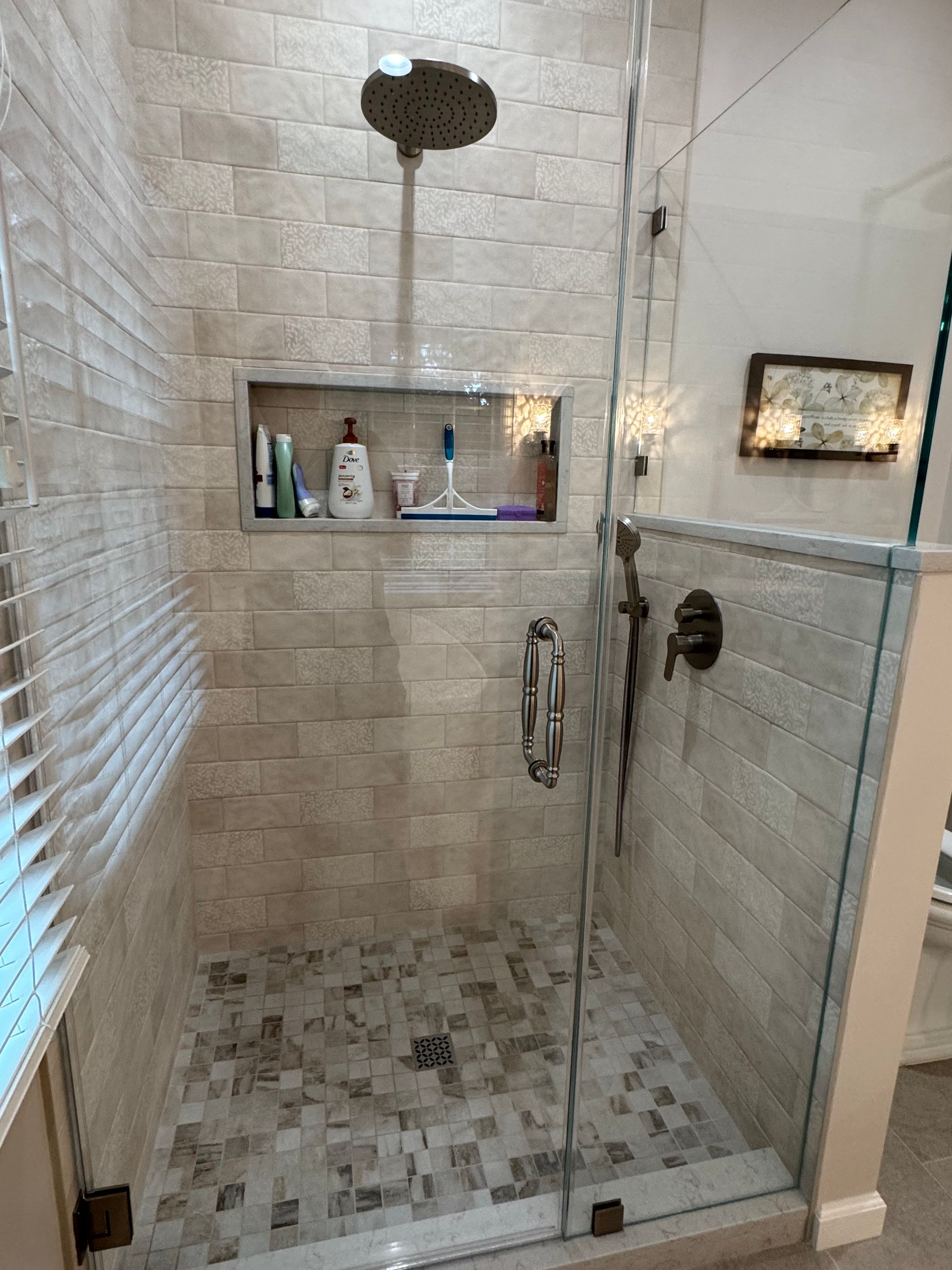 Modern walk-in shower with beige tile, rain showerhead, glass door, and built-in shelf with toiletries