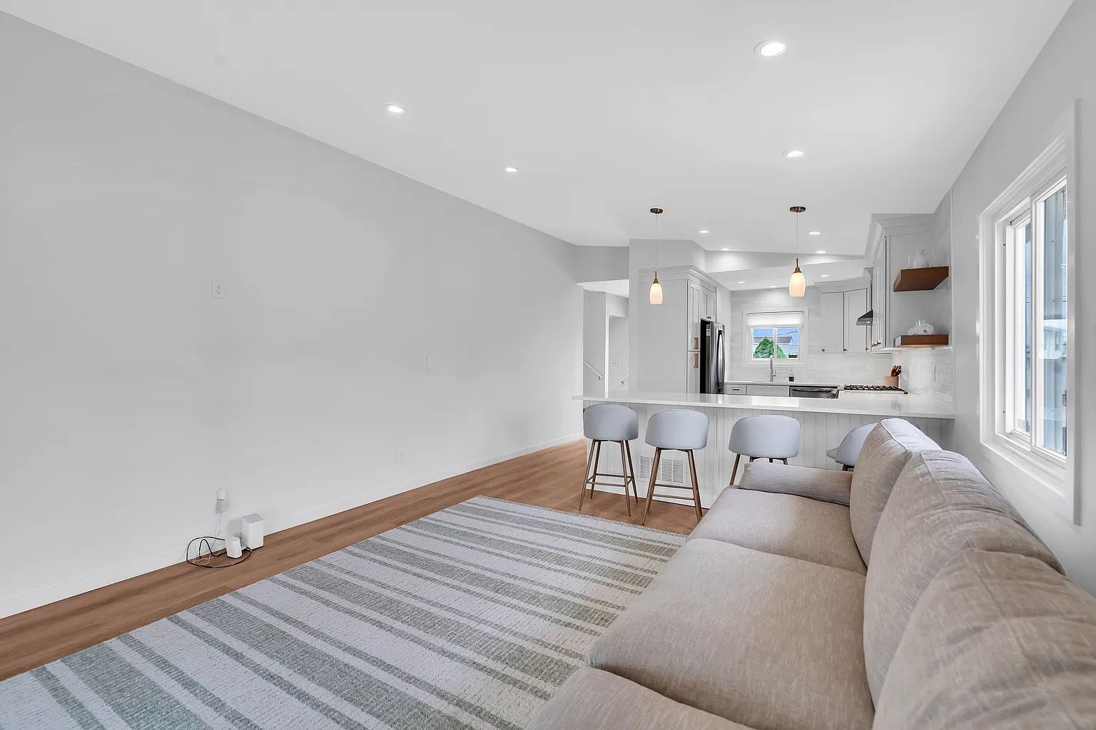 Bright open living room with beige sectional, striped rug, wood floor, and kitchen view in the background