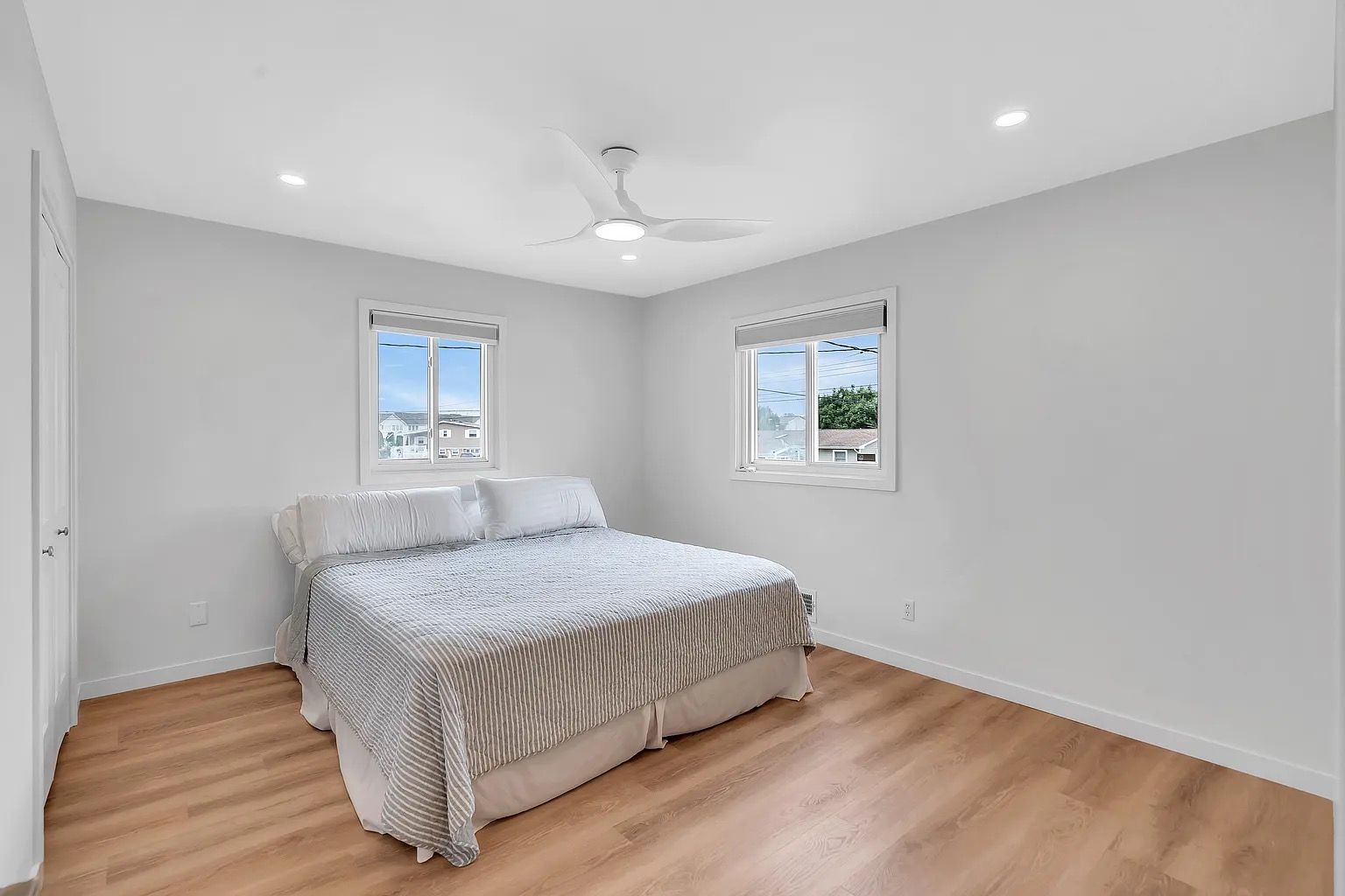 Spacious empty bedroom with white walls, light wood floors, and a bed beneath two windows