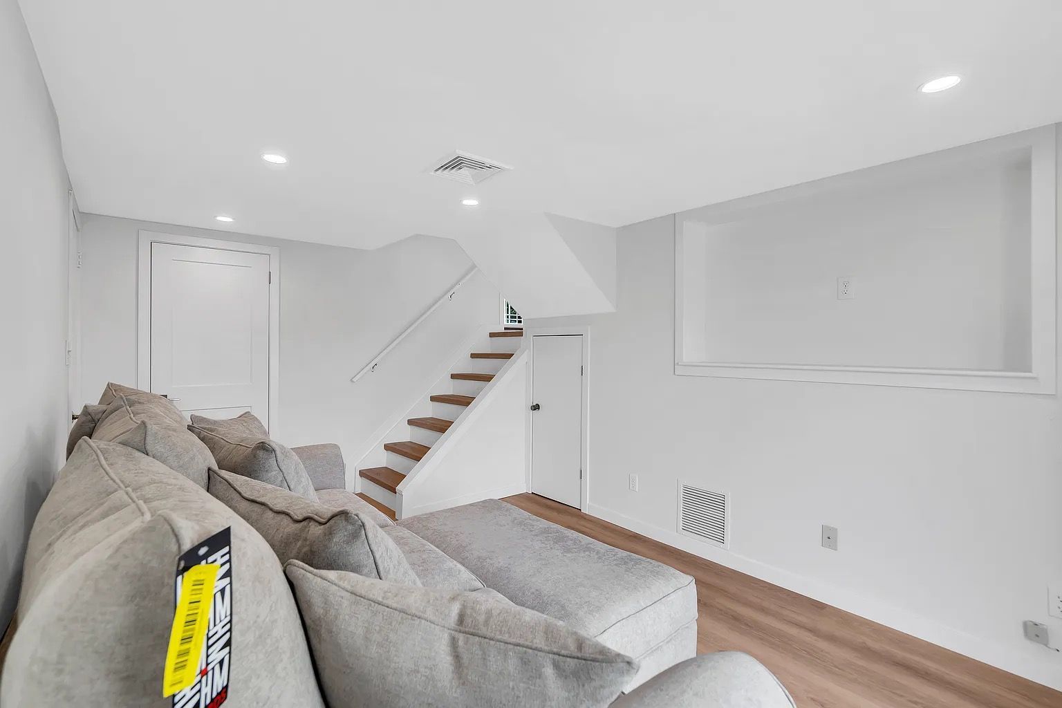 Bright modern living room with gray sectional, white walls, wood floor, and staircase