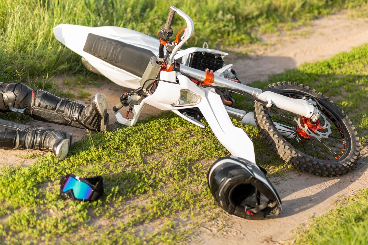 White dirt bike crashed on grassy ground, helmet, knee pads nearby. Motorcycle Accidents Law Attorney in Nashville, Tennessee