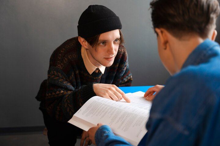 Person in beanie points to book held by another person. Dark sweater, blue shirt, gray wall. Second Degree Murder Law Attorney in Nashville, Tennessee