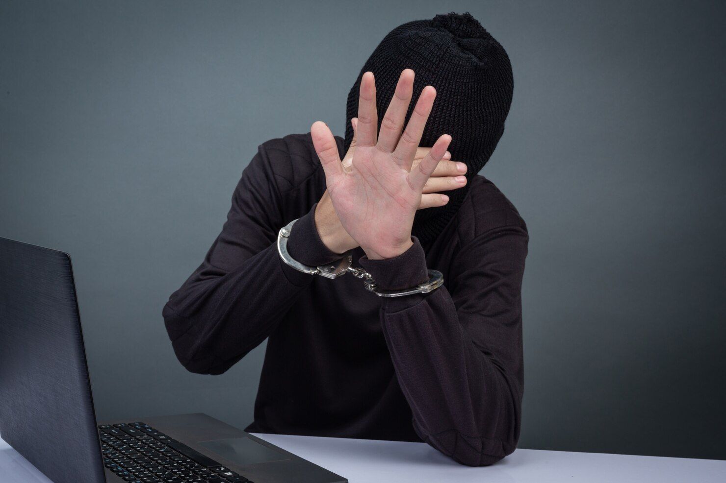 Person in a black balaclava and handcuffs, holding hands up near a laptop. Criminal Contempt Law Attorney in Nashville, Tennessee