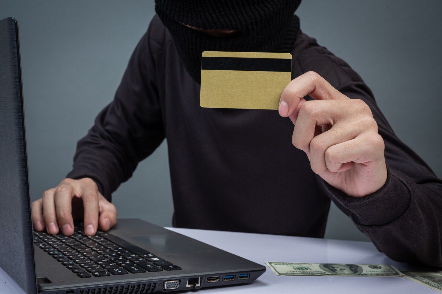 Person in black balaclava holds a gold credit card and uses a laptop with some money in the corner. Credit / Debit Card Fraud Law Attorney in Nashville, Tennessee