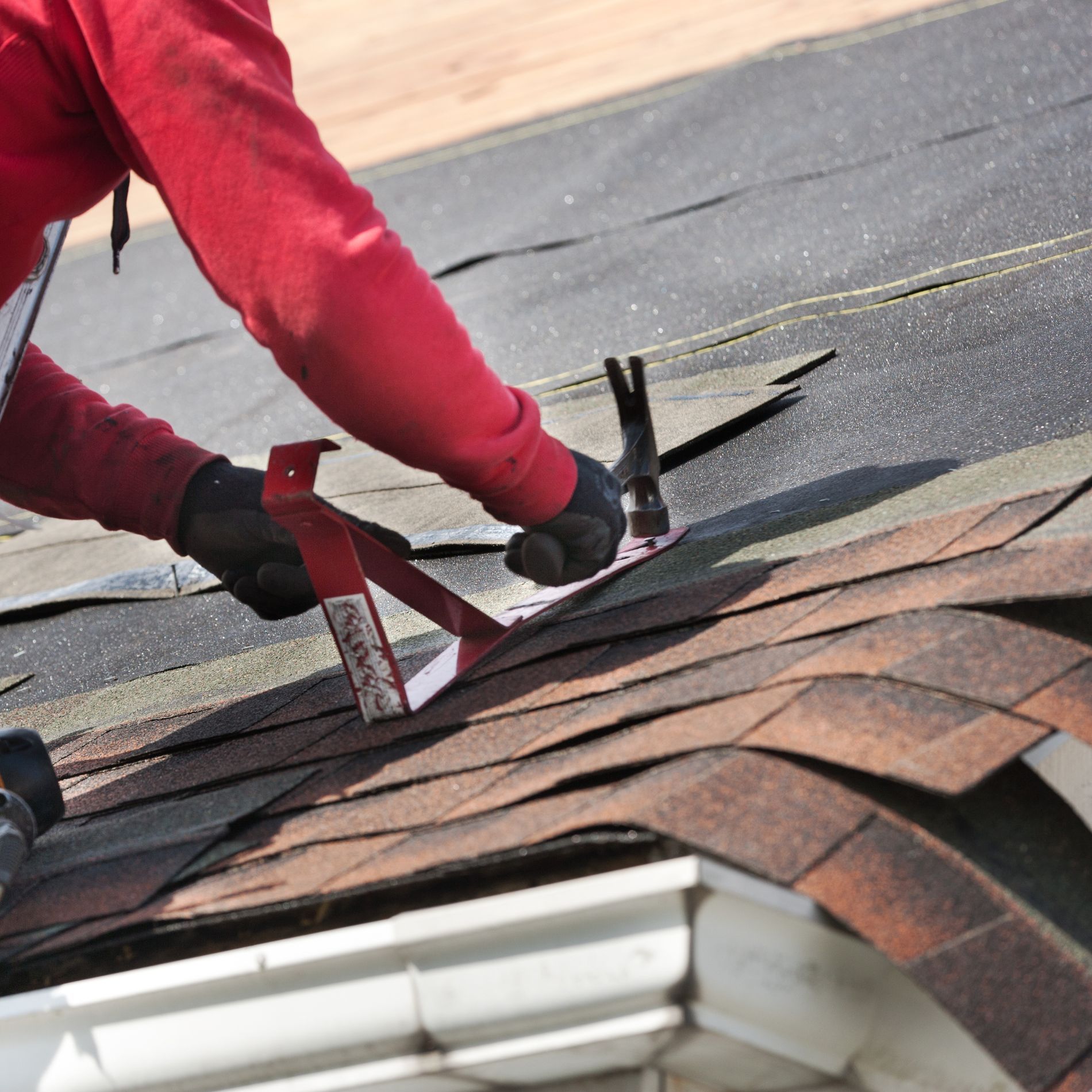 Roofer hammers roofing nails, using a guide, on a brown shingle roof.