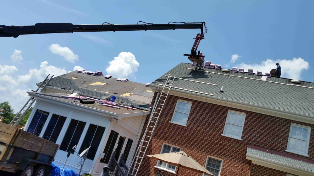 Roofing work in progress: Crane lifting materials to workers on rooftops of a brick house under a blue sky.